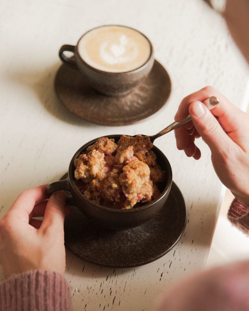 Person isst Apfel Crumble an einem weißen Tisch mit einem Cappuccino